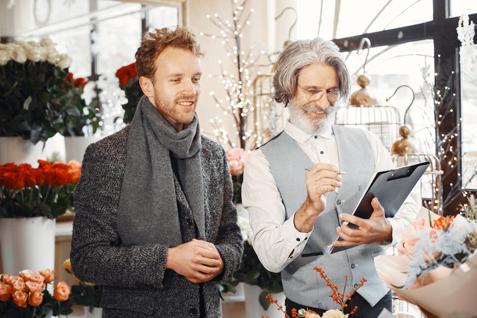 A male customer and a florist are inside a bright flower shop. The customer, a young man with curly hair and a grey scarf, is smiling and looking at the florist. The florist, an older man with long gr