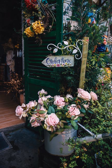 A floral arrangement featuring soft pink roses with lush green leaves, placed in a silver watering can, situated outdoors amidst various green plants and flowers. In the background, a dark green woode