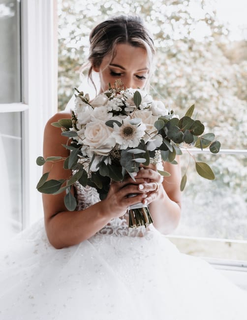 A bride holding a large floral bouquet featuring white roses, cream-colored anemones, and various eucalyptus leaves, with a mix of textures from smooth petals to matte foliage. The bouquet is wrapped 