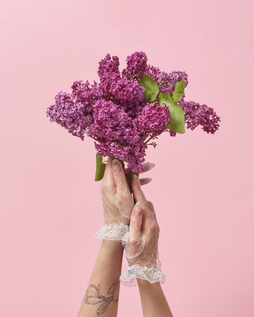 A person wearing delicate lace gloves holds a bouquet of fresh lilac flowers with vibrant purple clusters and green leaves against a soft pink background. The bouquet is hand-tied, with the stems wrap