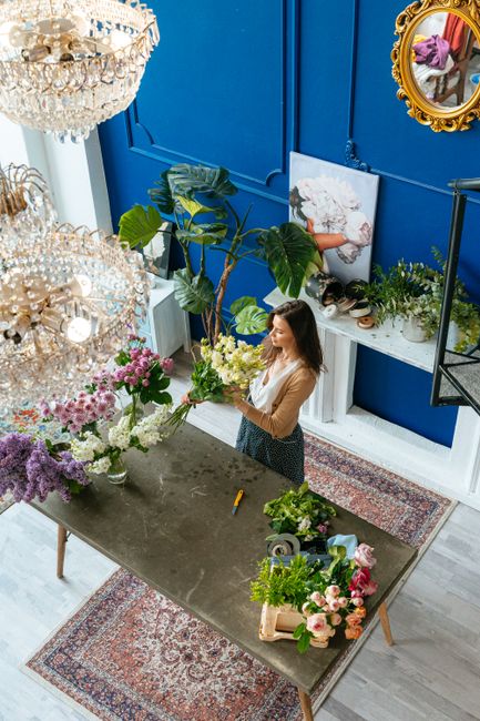 A woman with long brown hair, dressed in a beige top and patterned skirt, arranging a lush bouquet of white and pale green flowers on a dark wooden table in a brightly lit shop. The shop has a vibrant