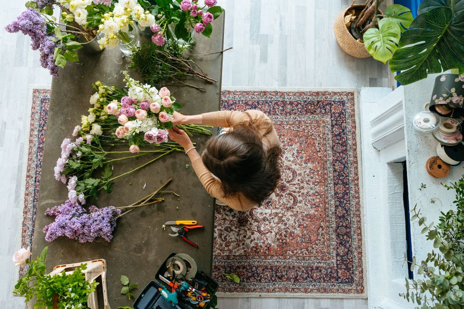 A florist arranging a bouquet of fresh flowers on a large, dark grey table in a bright, well-lit room. The bouquet includes pink and white roses, lilacs, and other seasonal blooms. The florist's hands